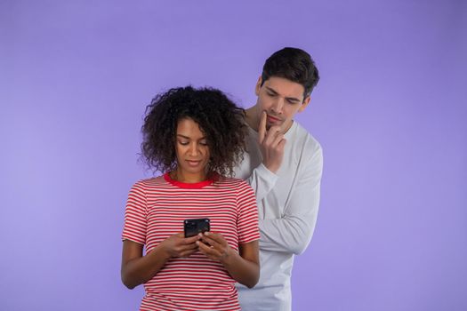 Handsome man is peeking over african girlfriends shoulder, she is communicating via mobile telephone.Guy shocked by jealousy and betrayal of wife.Purple background.Secrets concept, hidden double life.