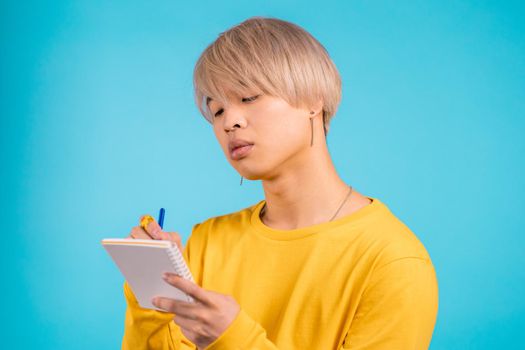 Asian man making notes in planner, guy holding pen. He writes future plans and to-do list in notebook for week or month. Keeping personal diary on blue studio background. High quality