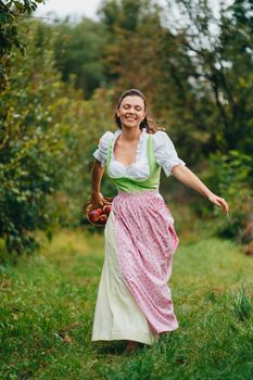 Happy woman in long retro dress with apron runs with basket in apple garden. Organic village lifestyle, agriculture, harvest, vintage style concept. High quality photo