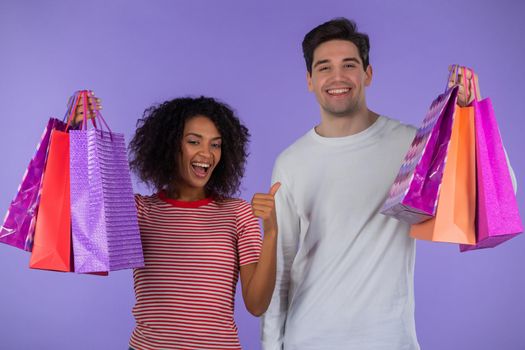 Interracial shopaholic couple holds shopping paper bags on purple studio background. White man and african woman bought presents on sales with discounts in center after quarantine. High quality photo