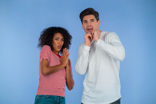 Frightened interracial couple afraid of something and looks into camera with big eyes full of horror on blue wall background. African american woman and white man. High quality