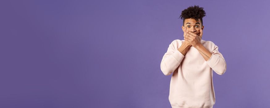 Portrait of shocked impressed young hispanic man found out gossip, heard rumour, trying not laugh shut his mouth with hands and express amazement and surprised with gaze, purple background.