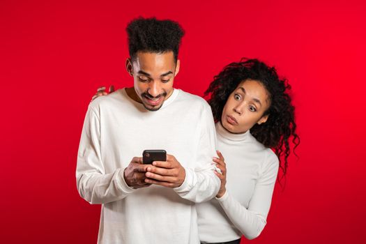 Pretty african girl is peeking over her boyfriends shoulder, he is communicating via mobile telephone. Woman shocked by jealousy and betrayal of her husband. Red studio background