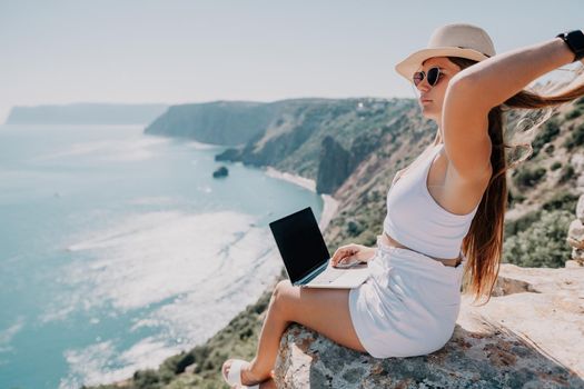 Successful business woman in yellow hat working on laptop by the sea. Pretty lady typing on computer at summer day outdoors. Freelance, travel and holidays concept.