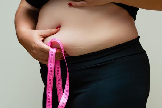 Cropped view of overweight woman measuring waist with tape measure at home, close up. Unrecognizable European woman checks result of weight loss diet or liposuction indoors.