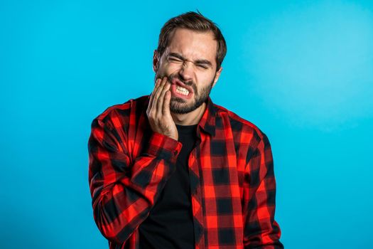 Young handsome man in red shirt with tooth pain on blue studio background. Toothache, dental problems, stomatology and medicine concept