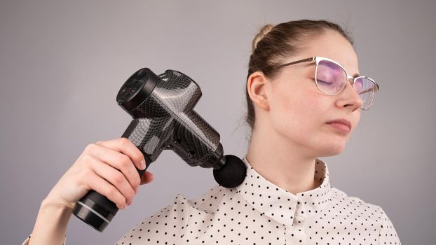 A caucasian woman uses a massager gun for pain in the muscles of her back and shoulders