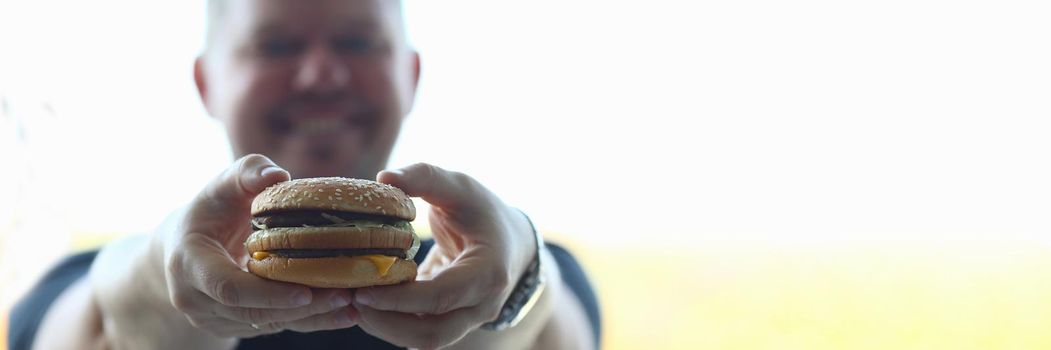 Young man holds hearty hamburger in hands. Fast food benefit and harm concept