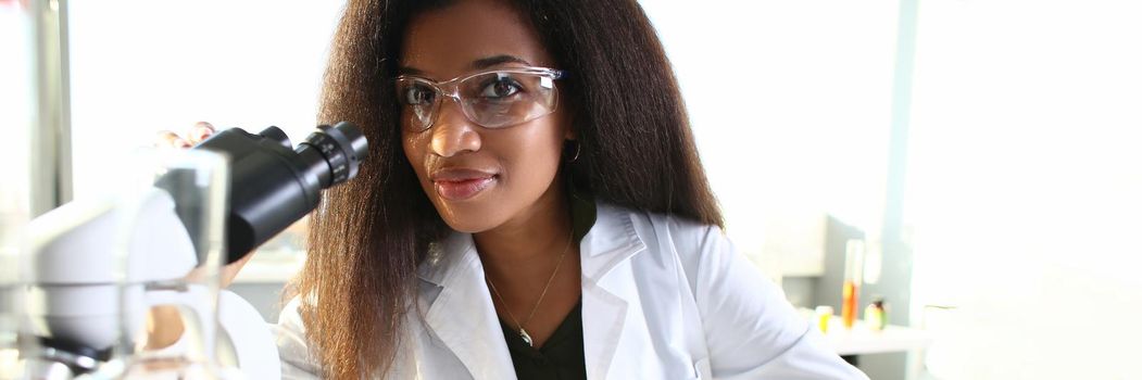 Black woman scientist in goggles, conducts study using microscope. Viral bacteria research concept