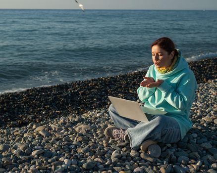 Caucasian woman talking by video link on laptop while sitting on the beach