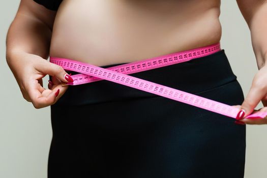 Cropped view of overweight woman measuring waist with tape measure at home, close up. Unrecognizable European woman checks result of weight loss diet or liposuction indoors.