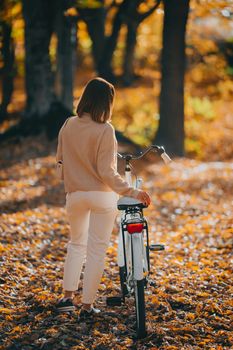 Beautiful portrait of woman cycling alone in autumn park. Sunny day, golden leaves in forest. Trendy lady on vintage white bicycle, healthy lifestyle, aesthetic scene. High quality photo