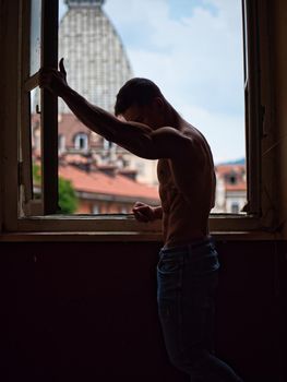 Portrait of sexy shirtless muscular man next to window curtains during the day, wearing only jeans, looking out