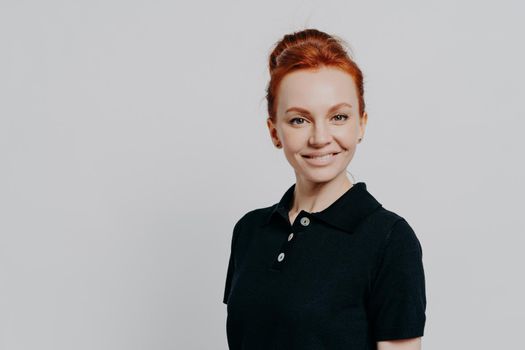 Gorgeous confident young 30s woman with red hair in bun posing isolated over grey studio background, dressed casually in black t-shirt, looking cheerfully at camera and expressing confidence