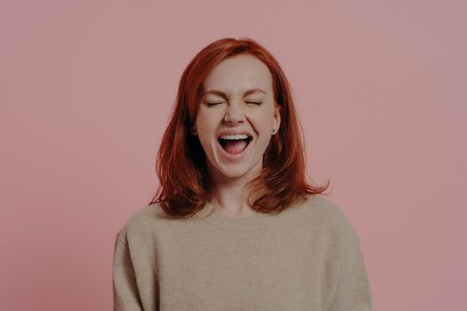 Overjoyed red haired caucasian woman laughing positively, with closed eyes, expressing positive emotions after hearing hilarious joke or anecdote, isolated on pink background. Face expressions concept