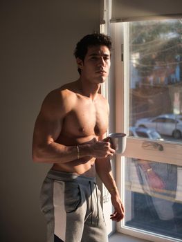 Sexy handsome young man standing shirtless in his bedroom next to window curtains, holding a coffee cup or mug, looking at camera