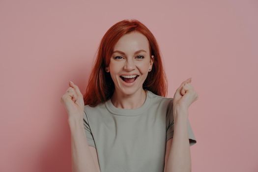 Young overjoyed redhead woman raising hands up with clenched fists, doing champion gesture, shouting yes with, winning prize while standing isolated over pink background.