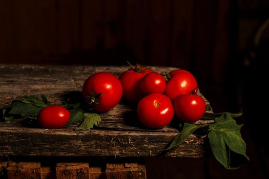 Red tomato fruits and green leaves on old wooden board on dark background.