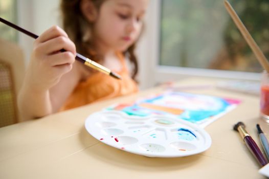 Focus on white plastic palette for mixing paints on the table on the blurred background of a little girl holding a paintbrush and painting with watercolour paints. Art master class. Child hobby