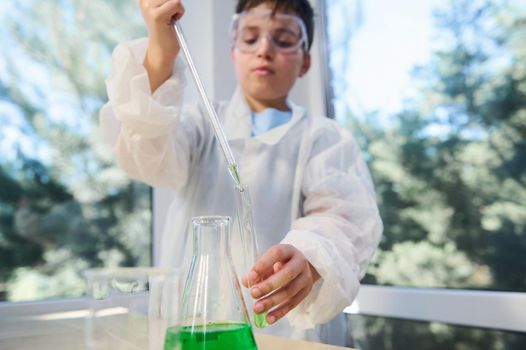 Details: Hands of a schoolboy holding a glass pipette and dripping few drops of green chemical solution from a flask into a test tube during a chemistry lesson in the school science laboratory