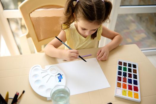 Top view of a little girl, learning watercolor painture, holding a paintbrush and painting with watercolor paints on a white blank paper sheet. Hobby and leisure. Kids entertainment and development