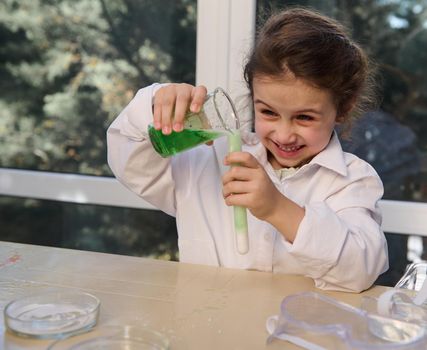 Cheerful little chemist, preschooler girl standing at the table and observing the chemical reaction going on in the glass flask with excited and surprised looks on her face. Miracles of chemistry