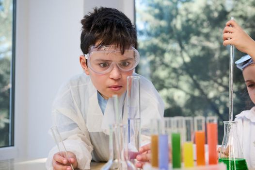 Curious schoolboy, little chemist in protective goggles and white lab coat, watching the chemical reaction taking place in test tubes with colored chemicals and reagents, at a chemistry lesson.