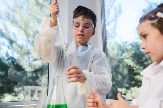Handsome Caucasian teenager, smart schoolboy in laboratory protective clothing, using a pipette, drips a reagent into a test tube, stands next to his classmate watching a going on chemical reaction