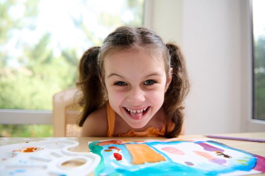Cheerful Caucasian little girl with two funny ponytails wearing an orange top, sitting at a table with her drawn picture, during a watercolor painting art class and grimacing looking at the camera