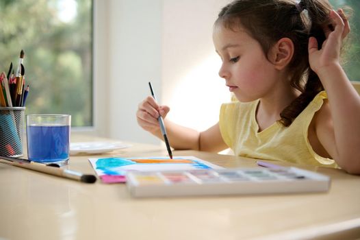 Beautiful cute child, adorable European little girl holding a paintbrush and drawing picture with watercolour paints. Art courses. Painting activity. Kids education and development concept