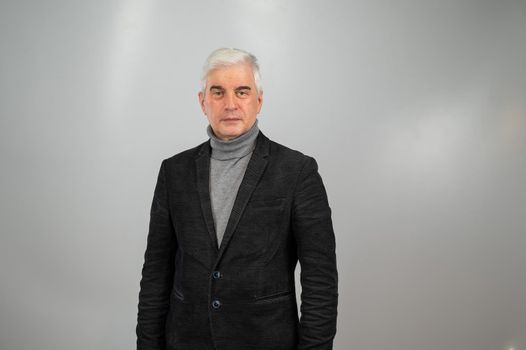 An elderly gray-haired man in a suit stands with his arms crossed over his chest