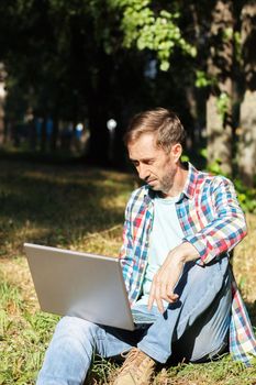 Adult man works on his computer in the park on the lawn.Remote work concept. The writer works remotely, enjoying nature.