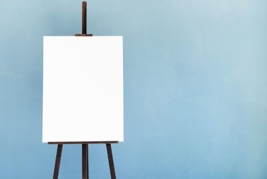 Wooden easel with blank canvas on light blue wall background for mockup design. Space for text