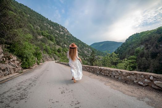 Female traveler in brown hat and white dress looking at amazing mountains and forest, wanderlust travel concept, atmospheric epic moment.