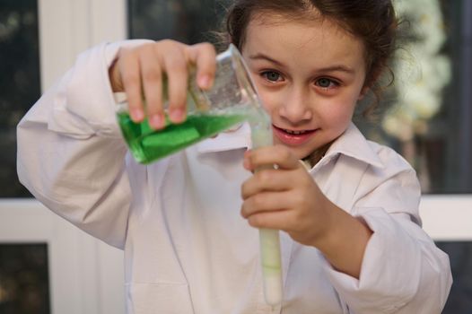 Homeschooling Caucasian little girl in white lab coat learning about science, pouring reagent solution from flask into a test tube, watching fascination going on chemical reaction. Learning chemistry