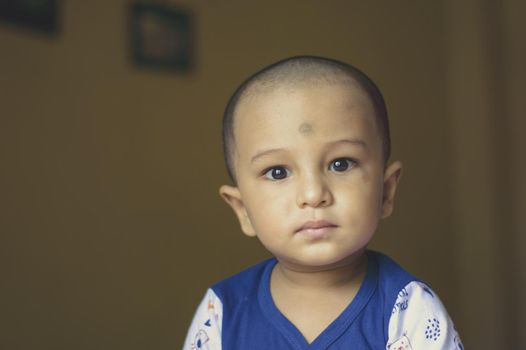 Cute bald indian baby boy in blue and white shirt looking away. Head and shoulder shot. Close up.