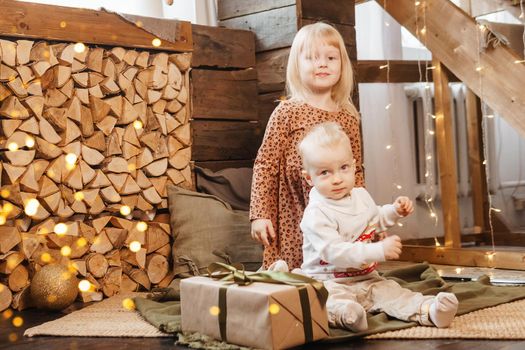 Little brother and sister play on Christmas eve in a beautiful house decorated for the New Year holidays. Children are playing with a Christmas gift. Scandinavian-style interior with live fir trees and a wooden staircase.