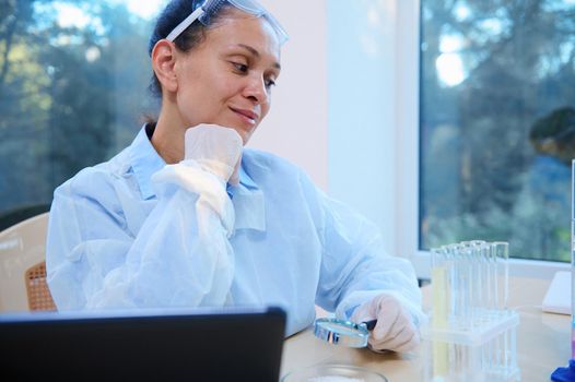 Confident experienced pleasant multi-ethnic woman scientist, health care worker, medical biologist, pharmacologist in white lab coat conducting scientific biological experiment in science laboratory