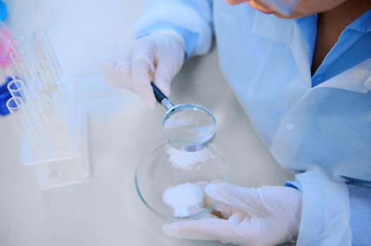 Cropped view scientist using loupe with magnifying glass to study pharmacological substance on Petri dish, sitting at table with empty test tubes on tripod in medical biolab. Investigation. Pharmacy