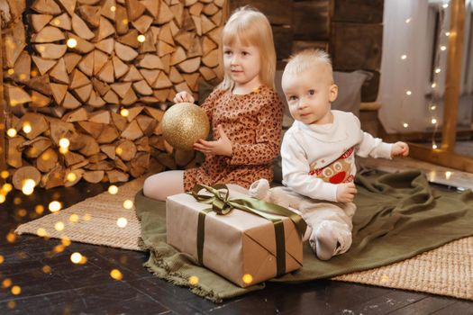 Little brother and sister play on Christmas eve in a beautiful house decorated for the New Year holidays. Children are playing with a Christmas gift. Scandinavian-style interior with live fir trees and a wooden staircase.