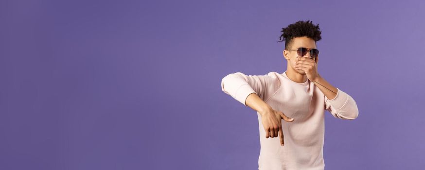 Waist-up portrait of cool young handsome guy with dreads, sunglasses on, hold hand over mouth while beatboxing, waving hand to shake audience up, reading own rap, purple background.