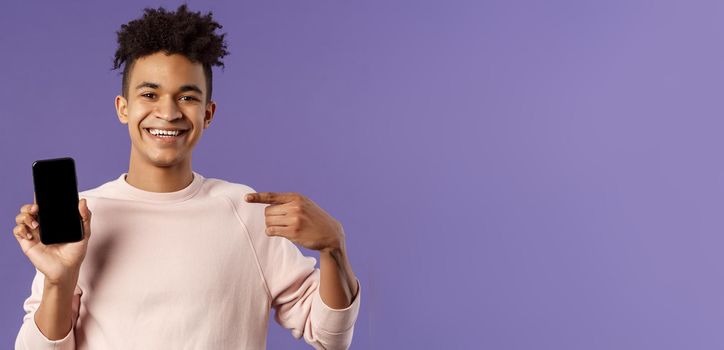 Close-up portrait of young handsome man promoting smartphone app or shopping online, internet delivery for goods, holding mobile phone pointing at display and smiling pleased, recommend.