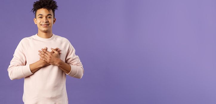 Portrait of romantic handsome hispanic man with dreads, hold hands pressed to heart and smiling pleased, see heartwarming nice gesture, receive lovely gift, being touched, purple background.