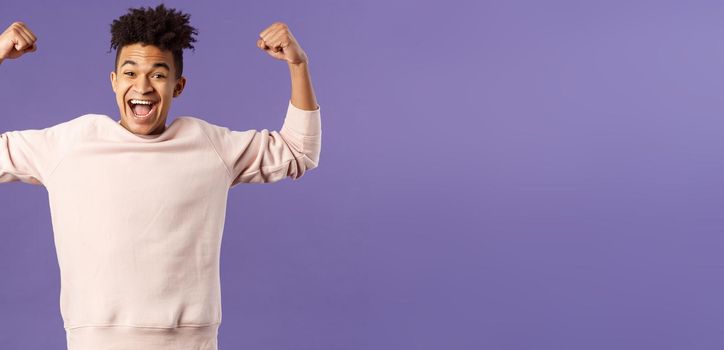 Portrait of young happy man got scholarship, applied to cool university, raise hands up flex biceps like champion, triumphing from great news, achieve goal and rejoicing, purple background.