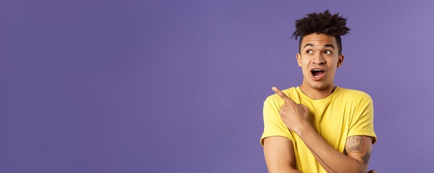 Portrait of impressed hispanic guy seeing something interesting, pointing finger and looking upper left corner, discuss strange thing upwards, standing purple background curious.