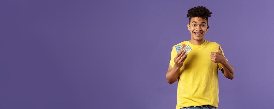 Health, influenza, covid-19 concept. Portrait of young healthy man got better, show thumbs-up holding drugs, taking pills to feel better after catching cold, being sick, purple background.