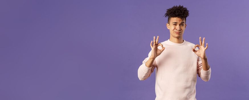 Portrait of skeptical young hispanic man rate something normal quality, show okay sign and grimacing judgemental, saying its not bad but average, standing purple background.