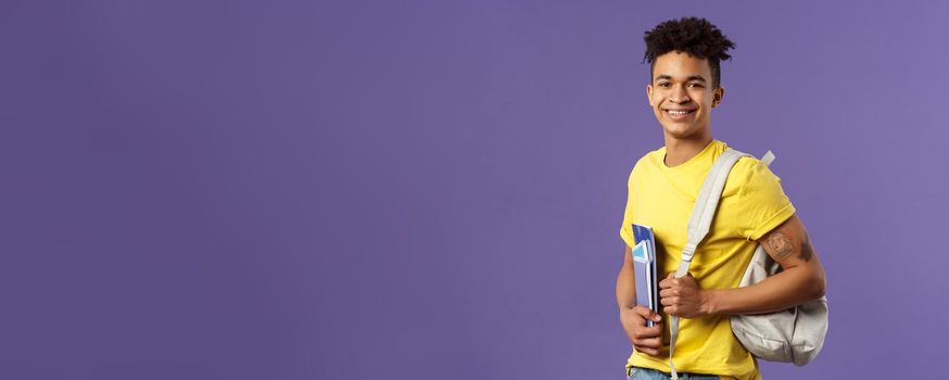 Back to school, university concept. Portrait of handsome charismatic hispanic guy, student walking to campus, going for lecture, studying hold backpack and notebooks, stand purple background.