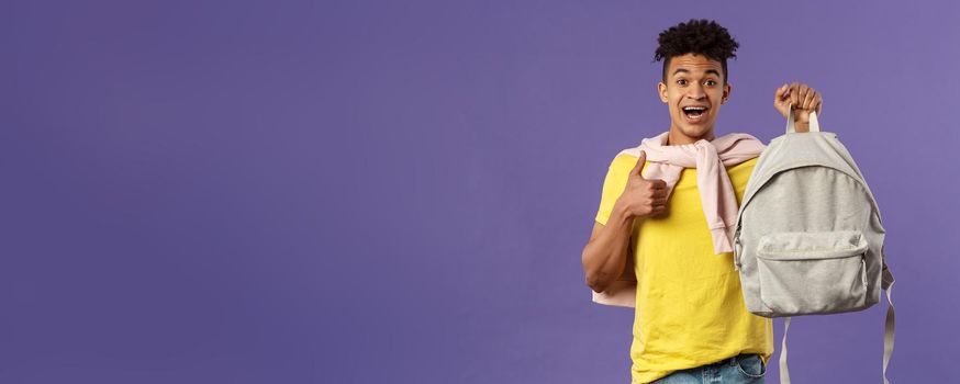 Portrait of cheerful young handsome male student, man recommending backpack, holding rucksack and show thumbs-up, bought new equipment for university semester, purple background.