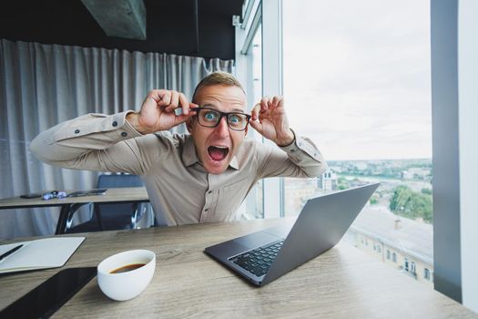 Emotional man looking at the camera at the workplace, a man in glasses, sitting at a desktop with a computer, a student is looking for inspiration for new ideas in an office cafe